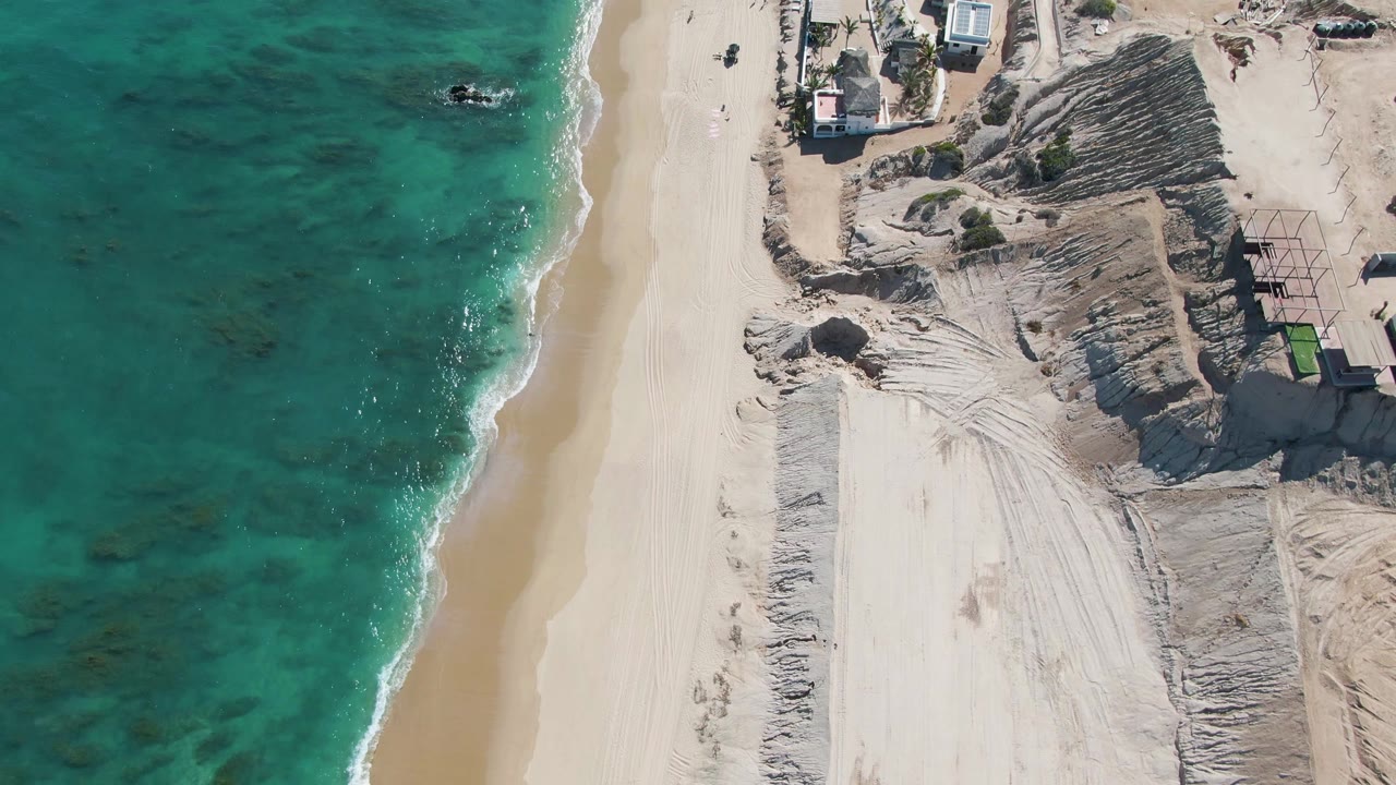 The La Fortuna aerial shows clear green-blue water, a narrow wave band, pale sand, rough dune grading, and vehicles spaced along the beach path, with no visible people.