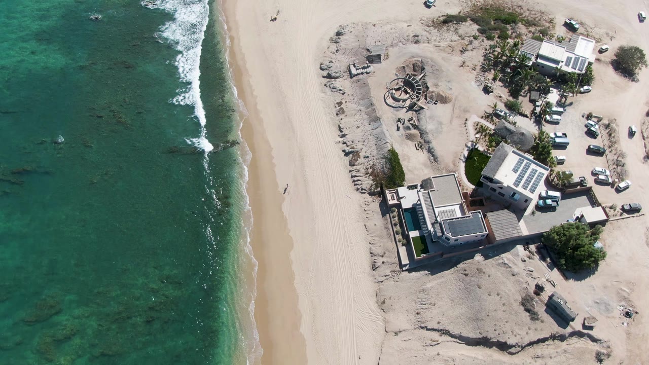 Overhead view in La Fortuna, Baja California Sur, with turquoise ocean and foam on the left, a broad sand beach, and beachfront buildings with solar panels and parked vehicles on the right.