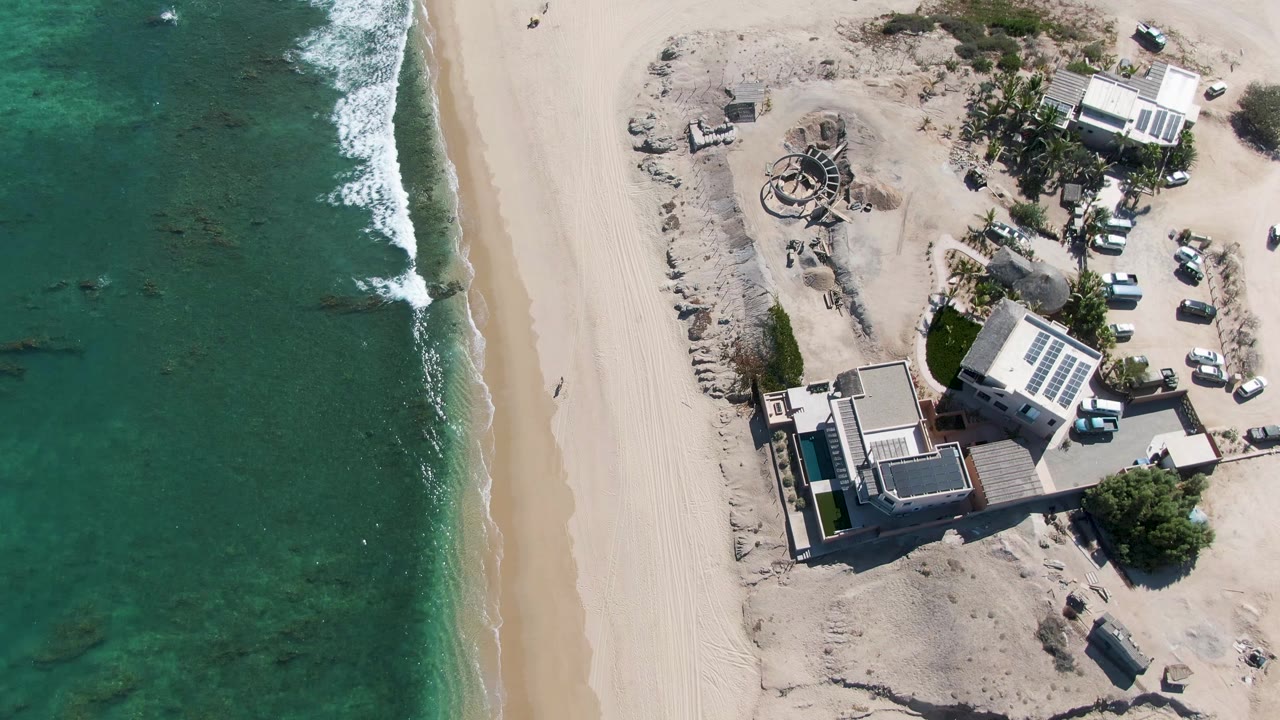 Overhead shoreline with waves beside a wide beach and nearby buildings.