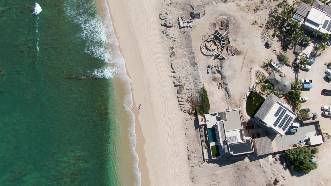 Aerial view over La Fortuna beach in Baja California Sur, with green-turquoise surf along pale sand, beachfront buildings with solar panels, palms, a white car, and a small visible person.