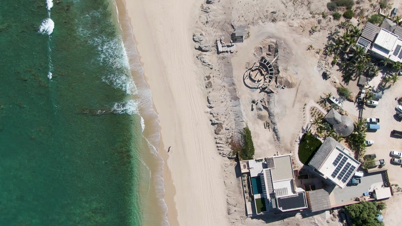 Vertical drone view of surf and sand beside beach buildings, palms, a white car, and a distant person.