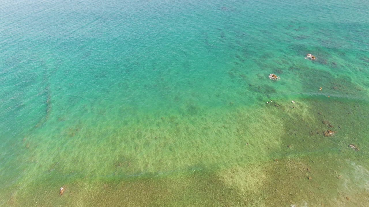 Aerial frame over La Fortuna shallow water, showing a broad green seabed area, turquoise surface ripples, and darker submerged shapes, no visible people.