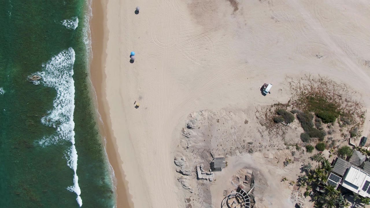 High overhead view of La Fortuna beach with green ocean and white surf along the left edge, pale sand through the middle, dune scrub, and small beachfront structures near the lower right.
