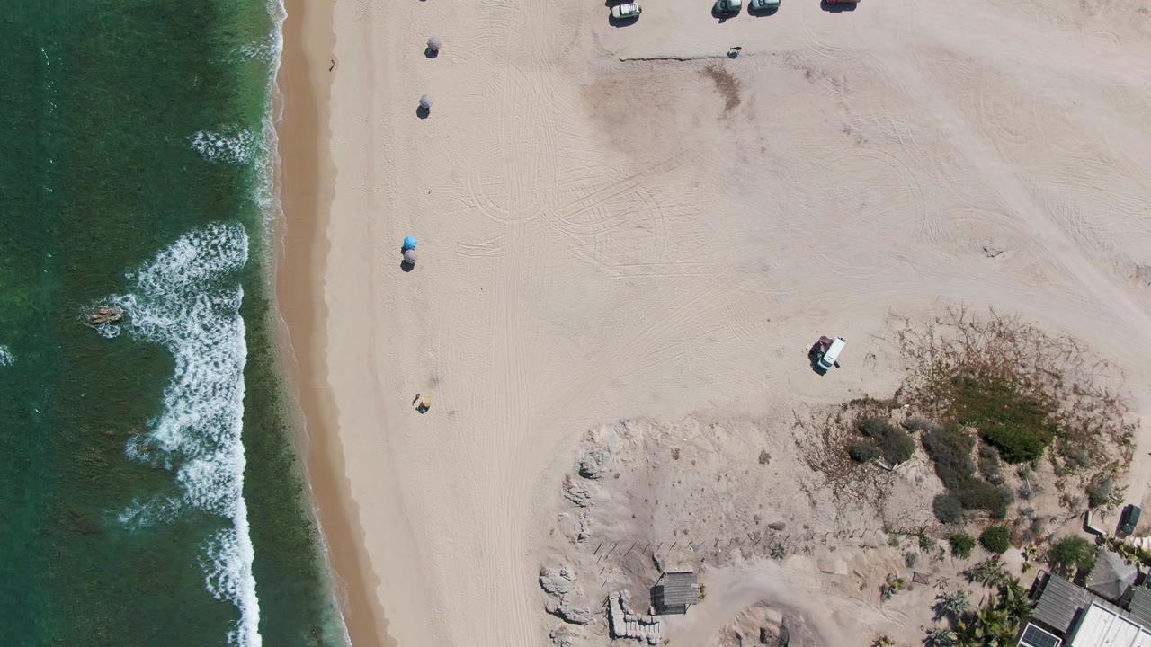Aerial beach view with surf, umbrellas, sand, and dune vegetation.