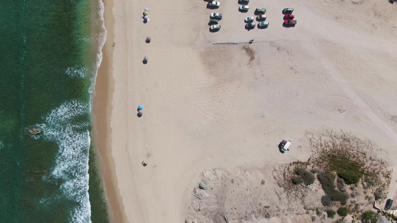 High aerial view over La Fortuna beach with more parked vehicles near the upper sand, small umbrellas in a line, green ocean at left, and scrub-covered dunes beside beach structures.