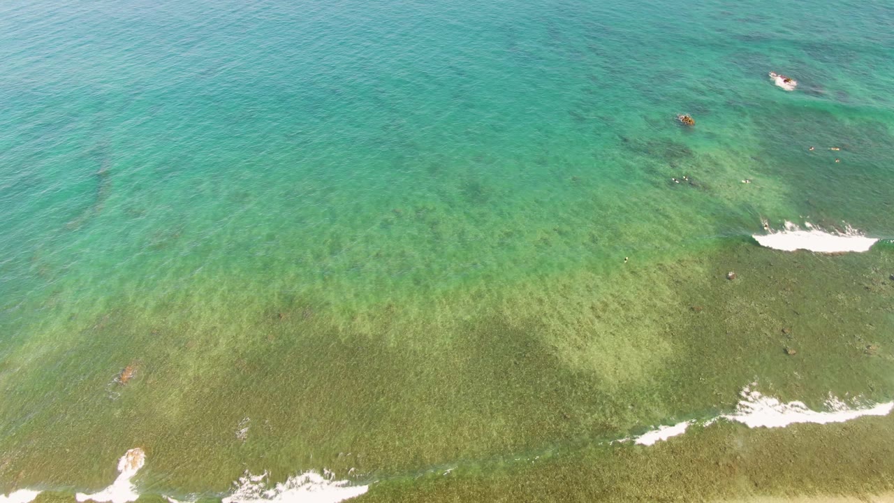Top-down aerial view of turquoise water and white foamy waves approaching a sandy La Fortuna shoreline, with no visible people.