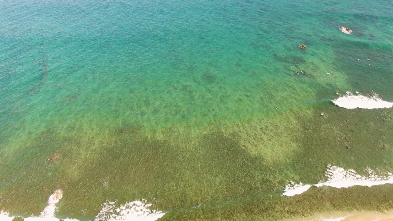 Aerial frame from La Fortuna with turquoise ocean filling the view, small white foam patches over shallow water, and pale sand starting to show at the bottom, with no visible people.