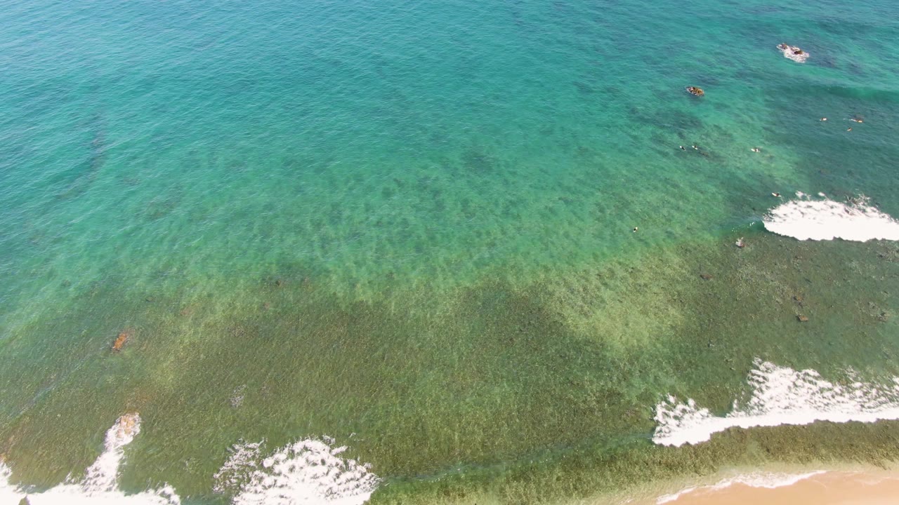 Near-vertical aerial view in La Fortuna showing teal water, a broad darker shallow band, white surf breaking near the lower right, and no visible people.