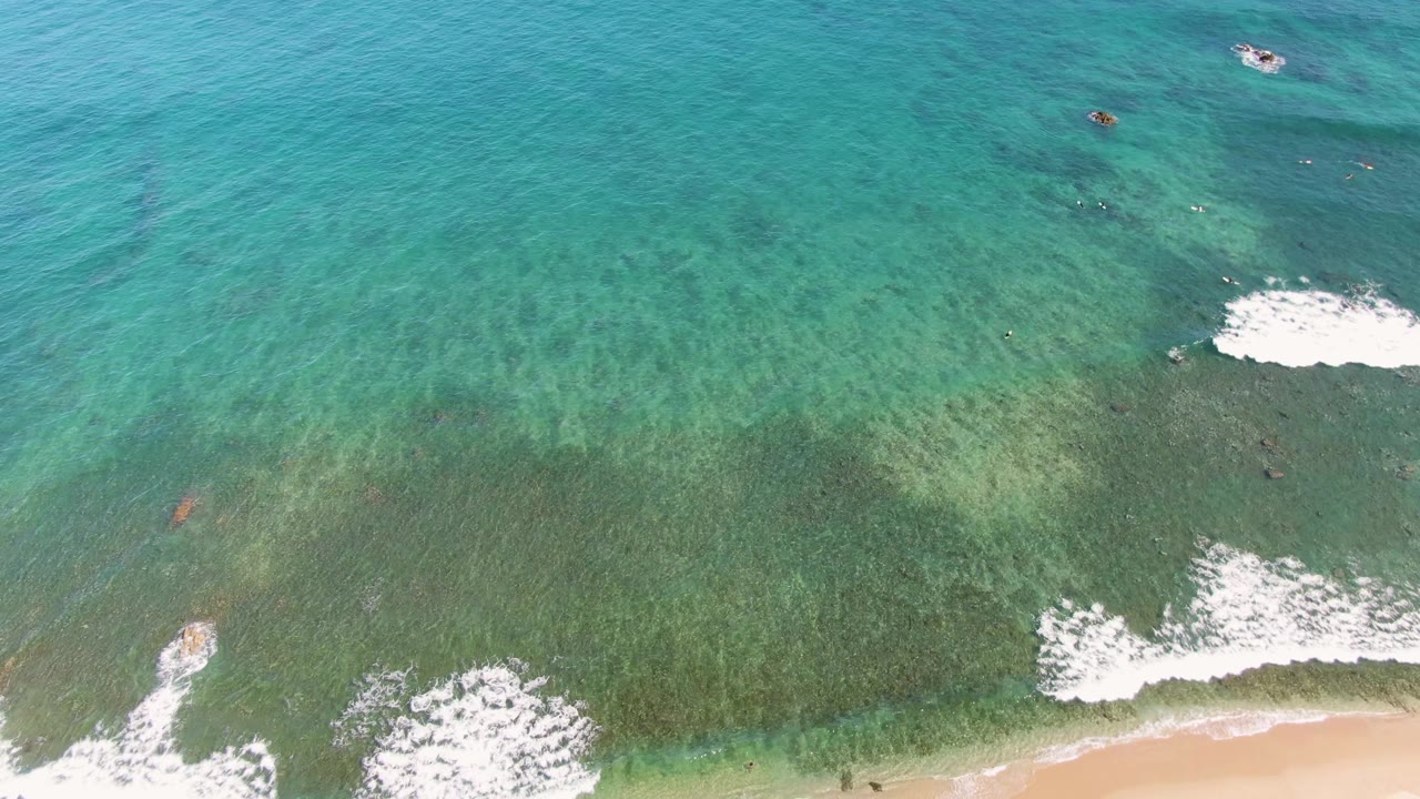Aerial ocean view with breaking foam and a sandy shoreline entering the frame.