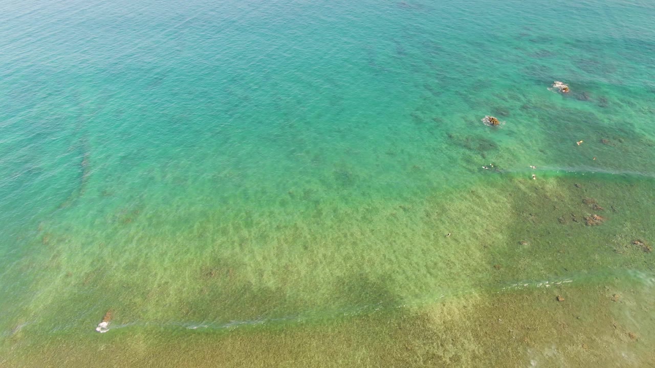Aerial view over La Fortuna, Baja California Sur showing turquoise shallow ocean water, green seabed texture, scattered darker patches, and small swimmers near the upper half of the frame.