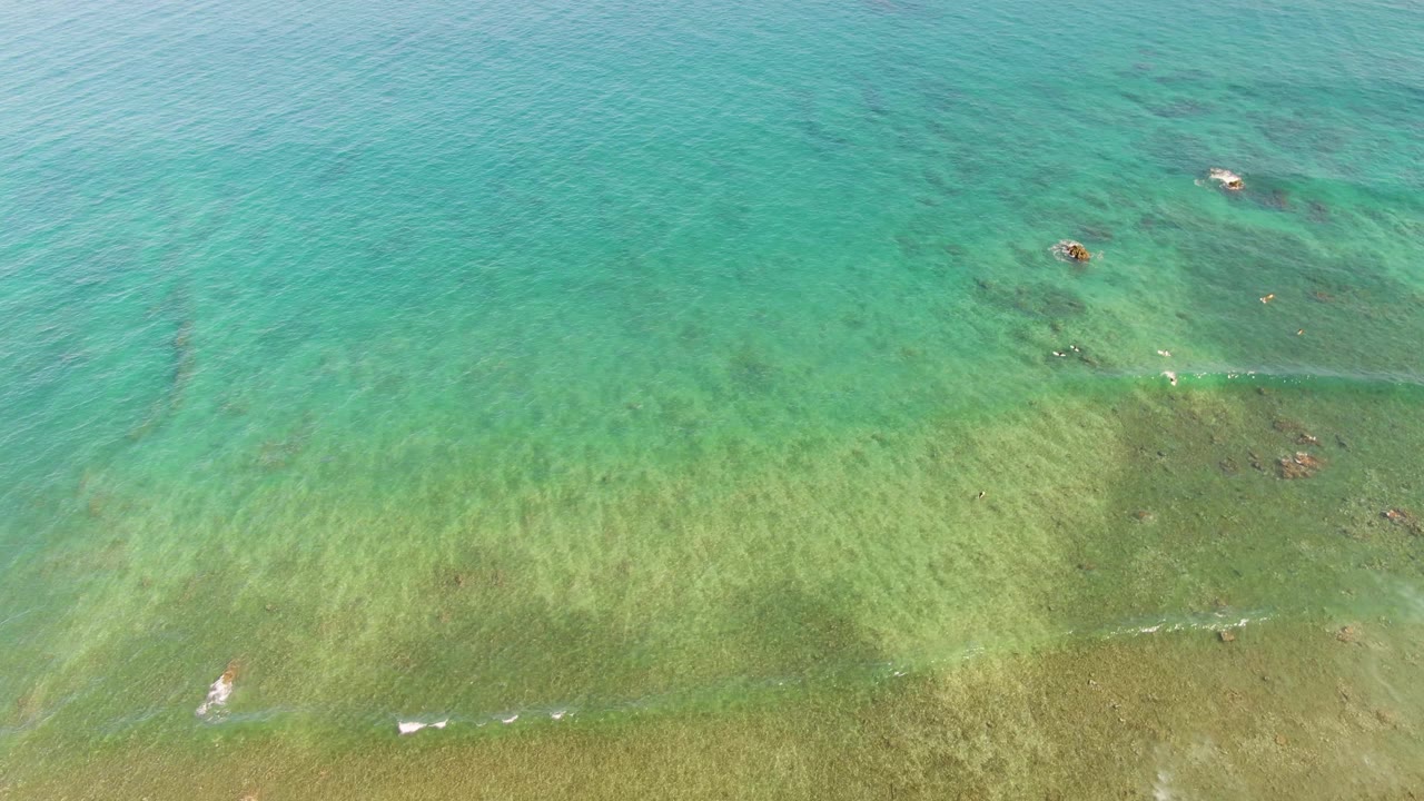 Clear turquoise water with seabed texture, small swimmers, and faint wave lines.
