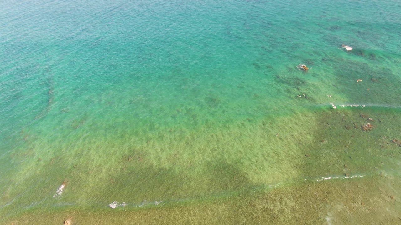 Steep aerial view over La Fortuna's shallow ocean water, with green seabed bands, turquoise surface ripples, tiny people in the upper water, and small dark reeflike patches.