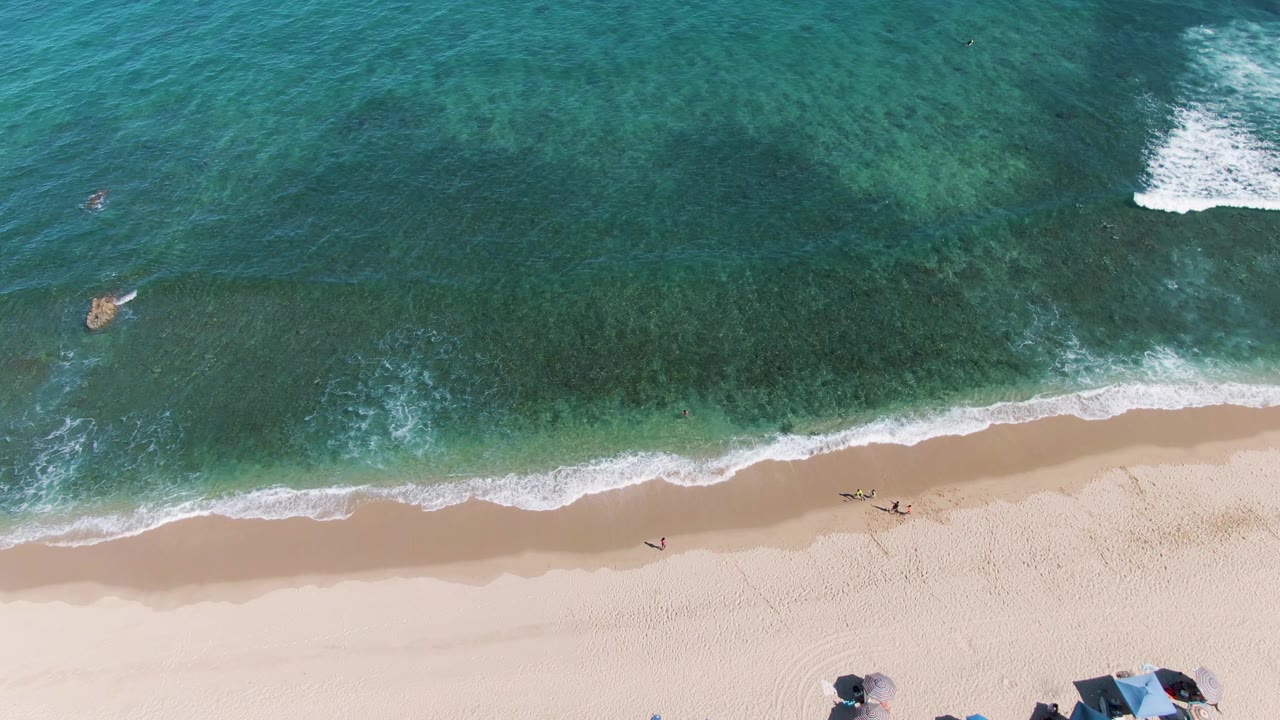 Overhead view of La Fortuna beach in Baja California Sur with turquoise water, a white breaking wave offshore, people near the surf, and umbrellas partly visible along the lower sand.