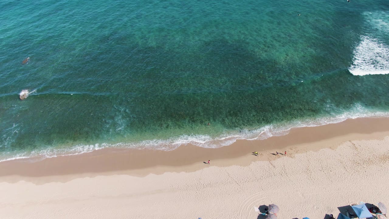 Top-down view of the La Fortuna shoreline with a broad band of turquoise water, foam curling onto pale sand, small figures walking and swimming, and umbrella tops clipped by the lower frame edge.