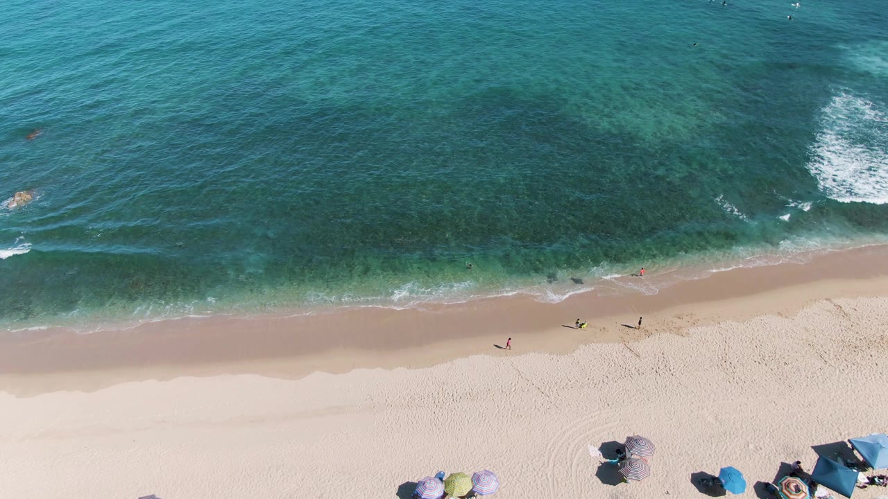 Overhead view of La Fortuna beach in Baja California Sur with turquoise water, a breaking wave, pale sand, scattered umbrellas, swimmers, and walkers near the shore.