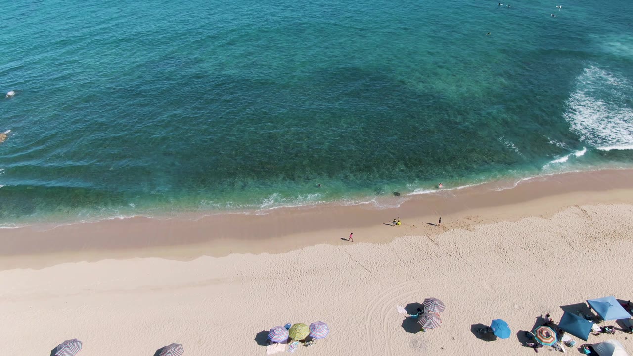 Aerial shoreline view with small waves, beach umbrellas, and people near the water.