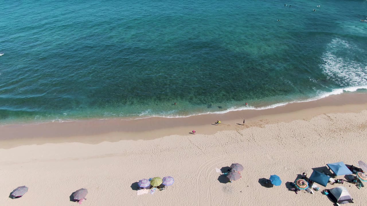 Drone view over La Fortuna beach with turquoise water across the top, a thin surf line, people walking along the water, and umbrella clusters along the lower sand.