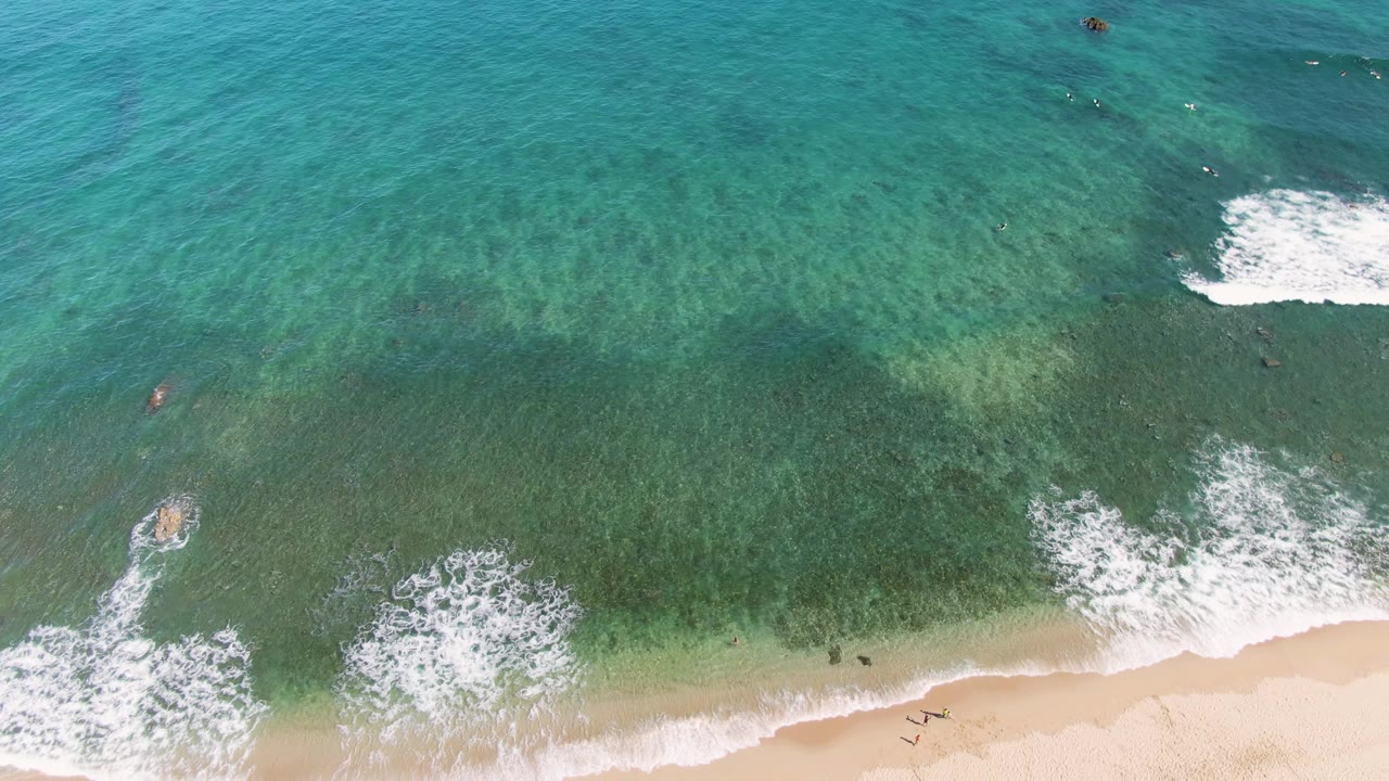 Overhead drone frame of La Fortuna beach in Baja California Sur, with turquoise water, white surf, pale sand, small swimmers offshore, and a few people walking near the shoreline.