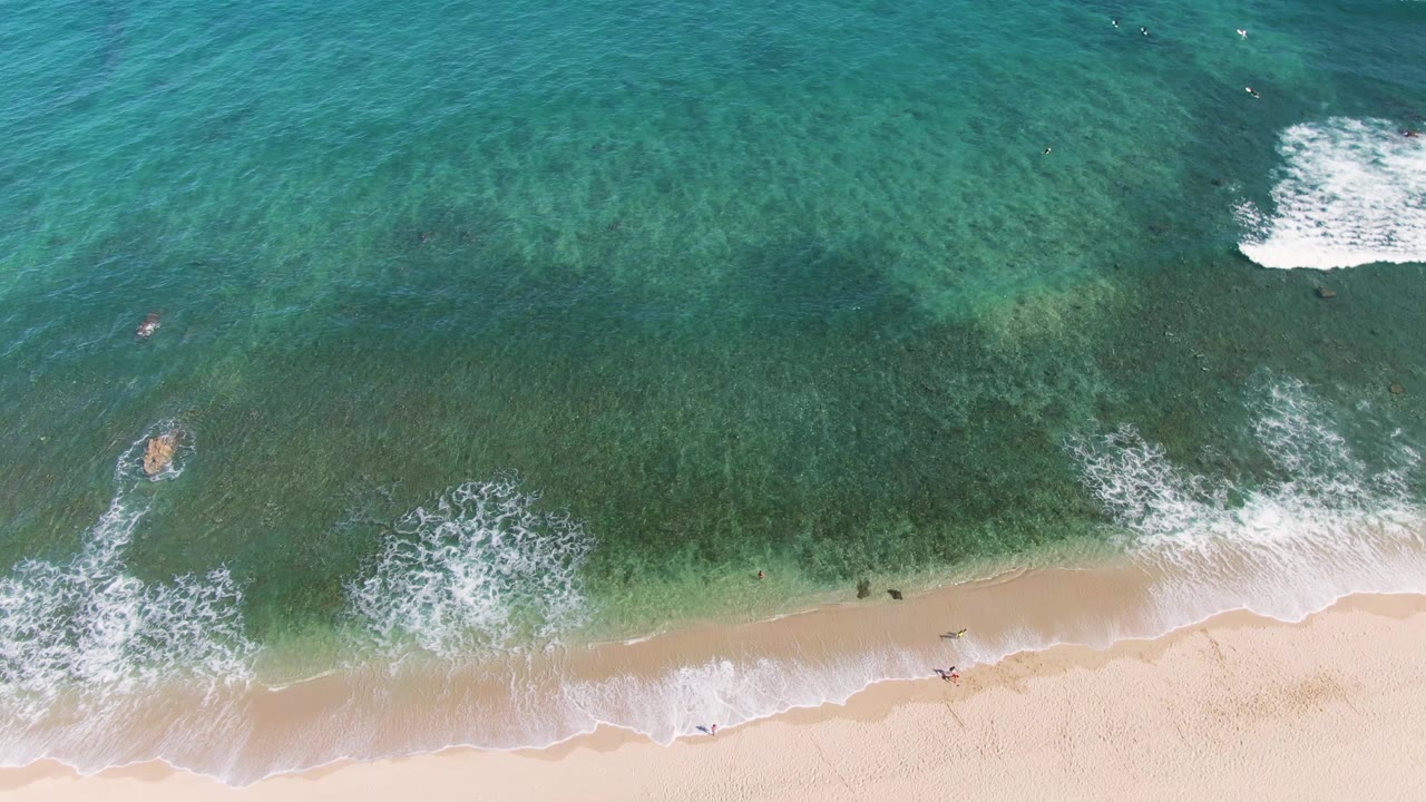 Overhead frame of the La Fortuna coast with foamy surf spreading toward the beach, darker green water beyond, a small swimmer offshore, and people spaced along the wet sand.