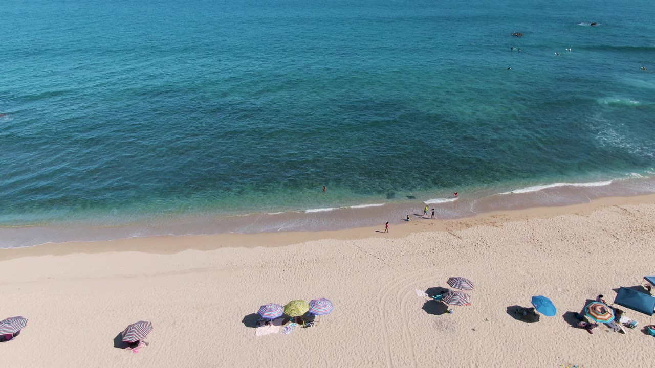 Aerial frame over La Fortuna, Baja California Sur showing pale sand, grouped beach umbrellas, small beachgoers near the shoreline, and turquoise ocean water under clear sky.