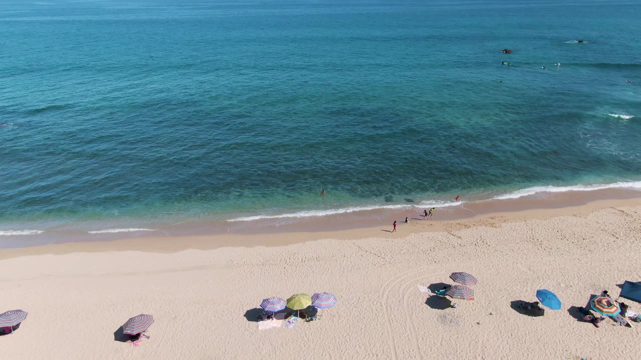 High aerial beach view with walkers near the surf and umbrellas on sand.