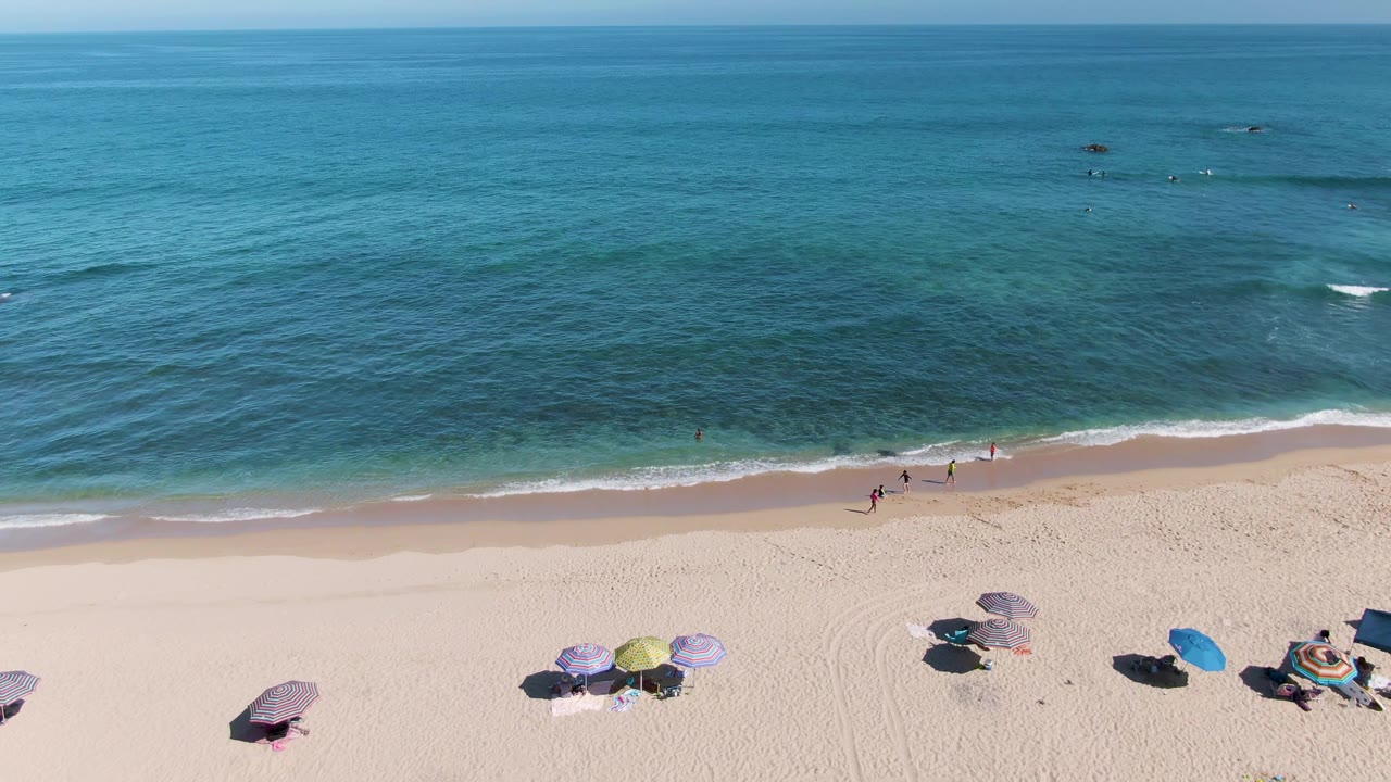 A drone-height frame over La Fortuna, Baja California Sur showing more open ocean above the shore, scattered swimmers offshore, beachgoers near the surf, and umbrellas along the lower sand.