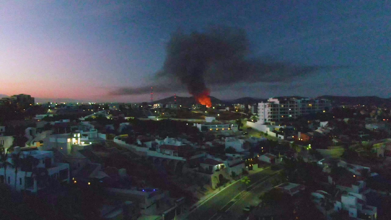 Aerial view over San Jose Del Cabo at dusk, with orange flames in the midground, a black smoke plume spreading across the sky, residential streets below, and no visible people.
