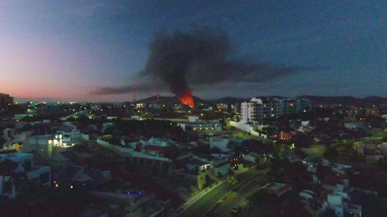 Aerial dusk frame in San Jose Del Cabo with the fire glowing near the center distance, smoke rising upward and drifting right, lit streets below, and no visible people.
