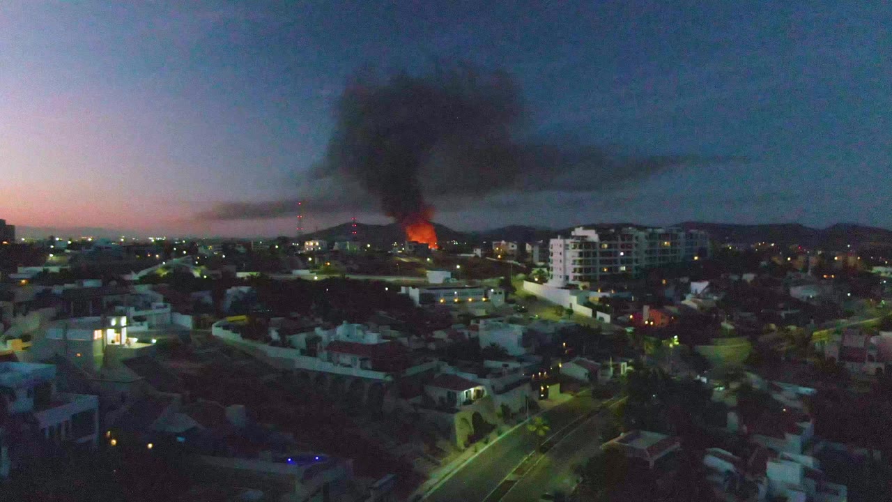 San Jose Del Cabo city blocks appear under a blue dusk sky as a wide black smoke plume extends from the fire across the horizon, with no visible people in frame.