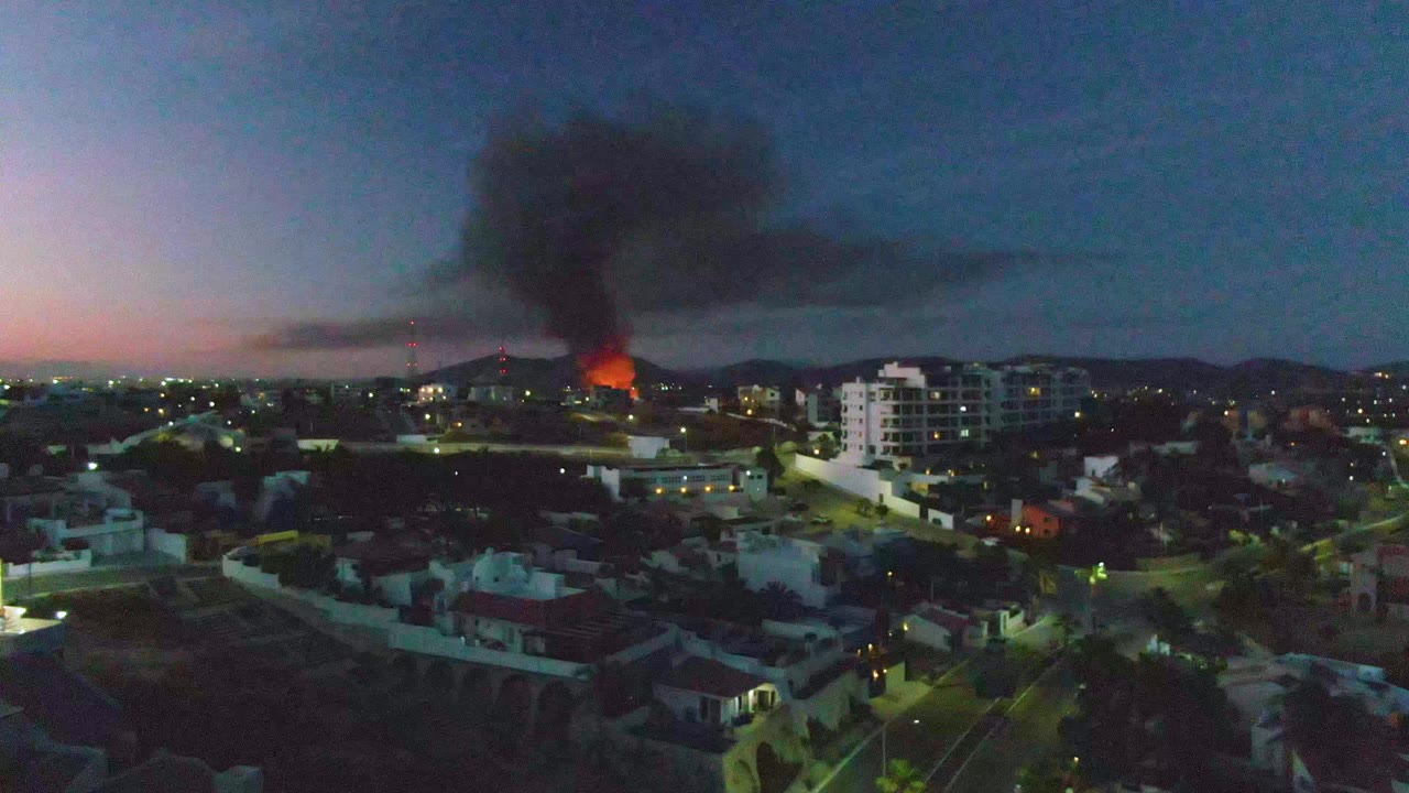 Aerial dusk view in San Jose del Cabo, Mexico, with orange flames in the midground, a dark smoke plume over low buildings, lit streets below, and no visible people.
