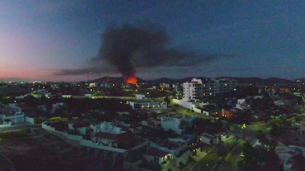 Urban dusk cityscape with orange flames and a heavy smoke plume near the horizon.