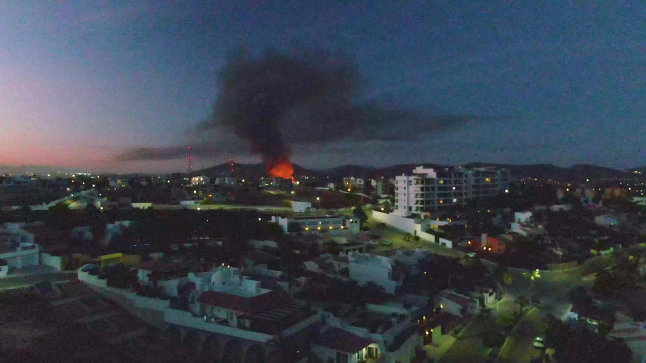 San Jose del Cabo city blocks sit under a darkening sky as orange flames burn in the midground and a broad black smoke plume drifts across the frame, with no visible people.