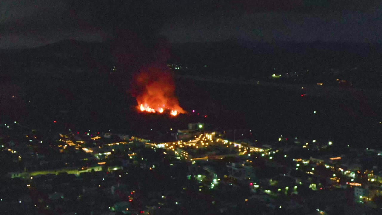 Night hillside fire above illuminated city lights.