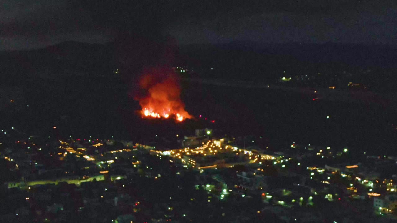 Aerial night frame of San Jose del Cabo showing a bright orange fire on the hillside and a cluster of street lights below, with no visible people.