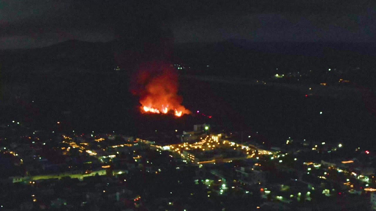 Aerial view over San Jose del Cabo with flames and smoke on the left side of the hillside and illuminated roads below, with no visible people.