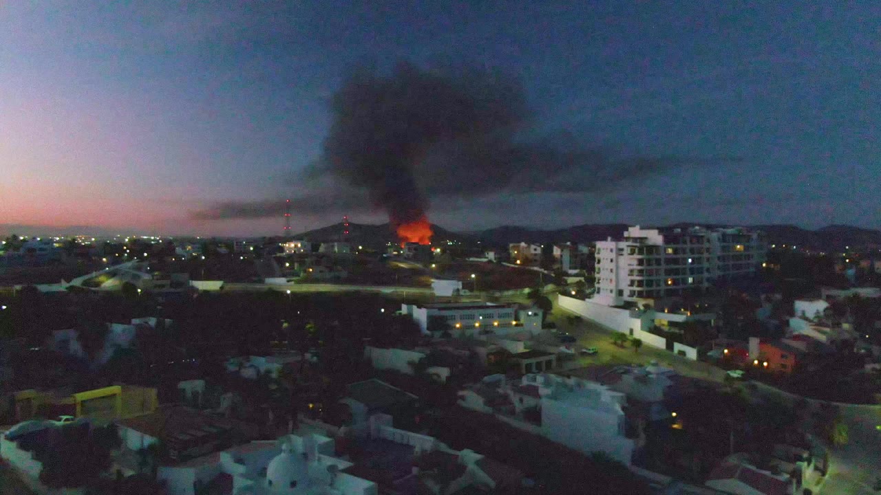 Wide elevated view in San Jose del Cabo showing a distant orange fire, black smoke rising over low buildings, lit streets, and no visible people at the start of the clip.