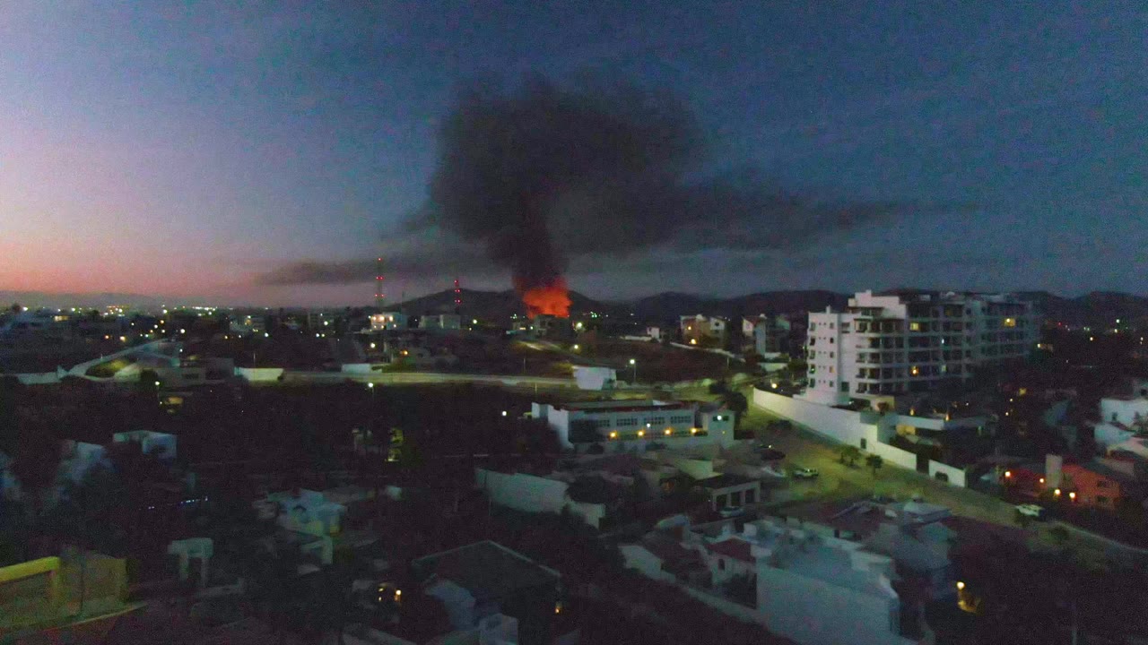 San Jose del Cabo aerial-style view with a bright fire beyond midground buildings, black smoke drifting across the upper frame, city lights, and no visible people.