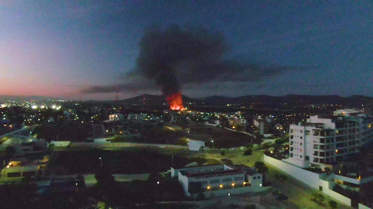 Distant orange fire sends black smoke above San Jose Del Cabo buildings and roads at dusk, with no visible people.