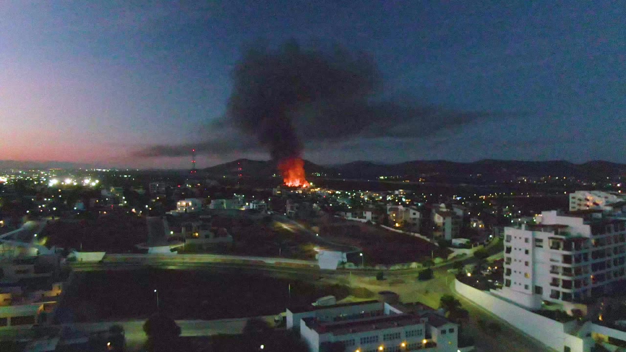 A thick smoke column spreads above San Jose Del Cabo while street lights and apartment blocks fill the lower frame, with no visible people.