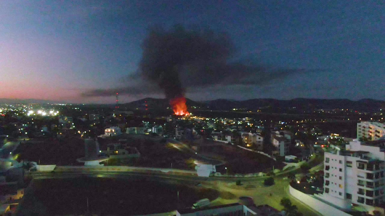 The fire burns near the horizon in San Jose Del Cabo as black smoke drifts across the upper sky over lit streets, with no visible people.