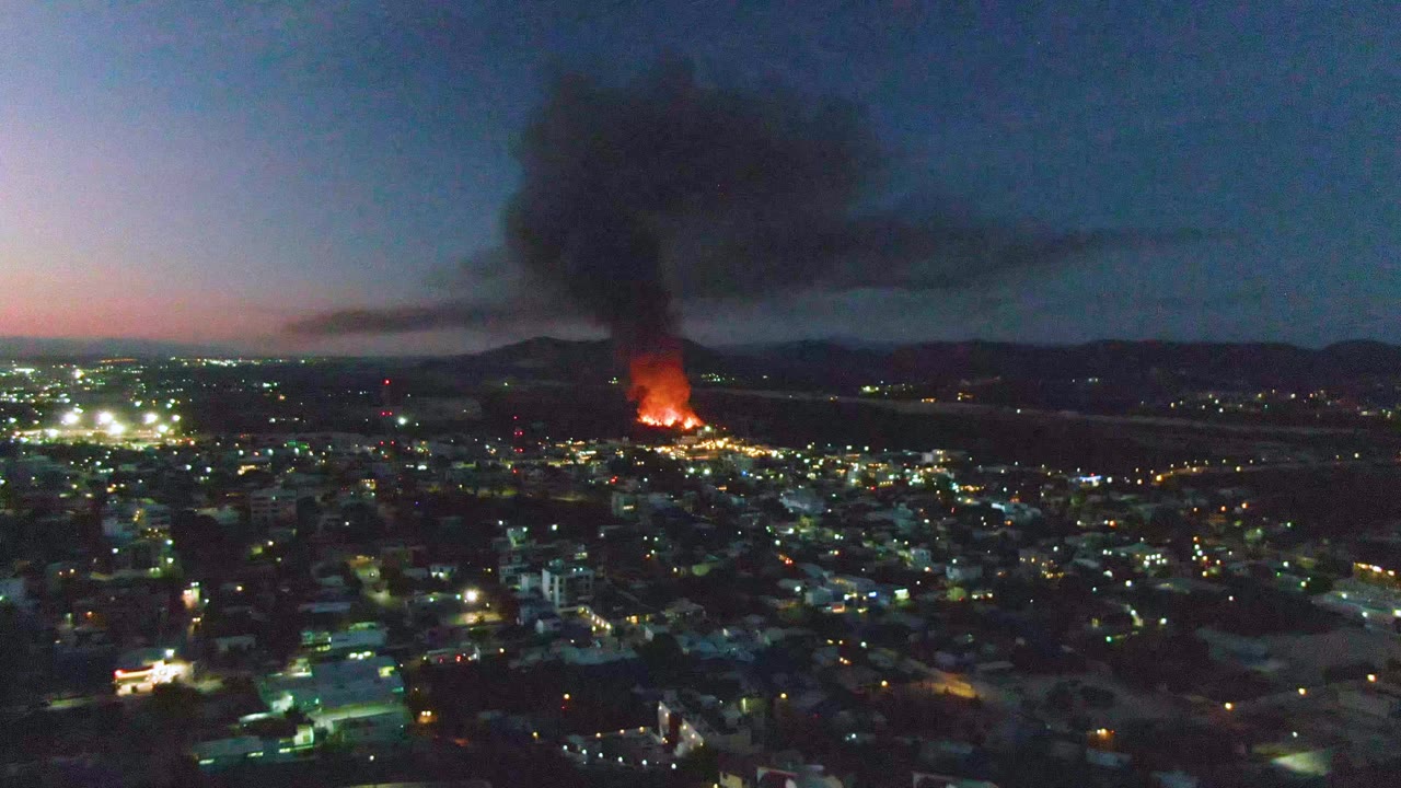Aerial dusk view over San Jose del Cabo with city lights below, orange flames in the distance, a black smoke plume rising, hills on the horizon, and no visible people.