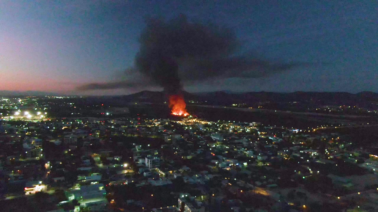 Aerial view across San Jose del Cabo at dusk, with bright city lights in the foreground, a fire burning near the horizon, heavy black smoke spreading upward, and no visible people.