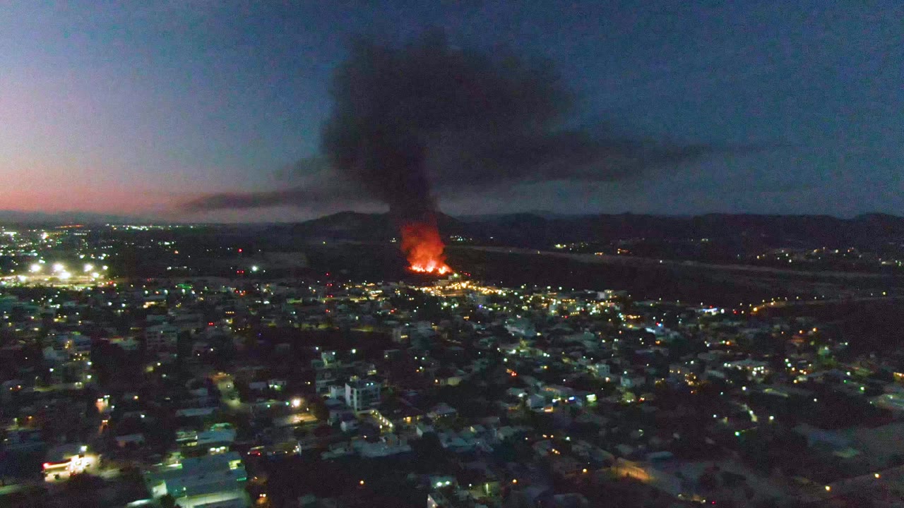 San Jose del Cabo city lights stretch across the foreground as orange flames burn in the midground and a dense black smoke column rises into the dark blue evening sky, with no visible people.