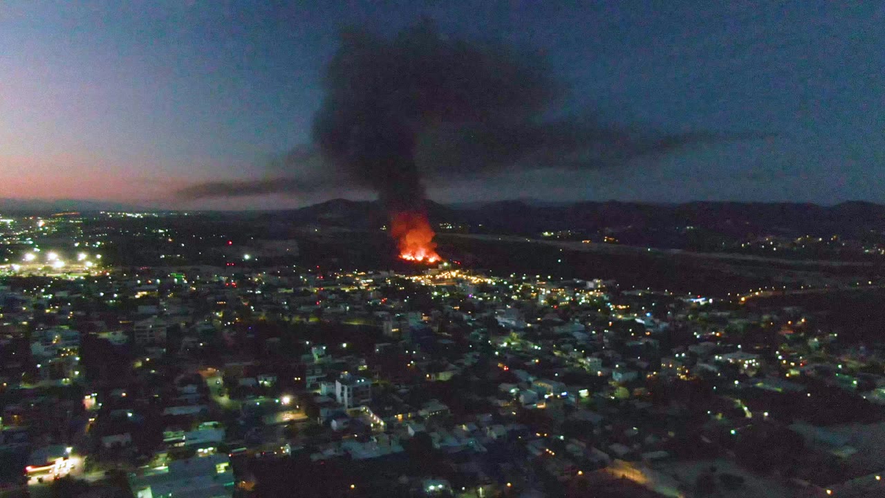Wide dusk aerial of an urban fire with smoke over a lit city.