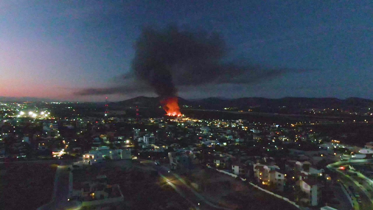 Aerial dusk view over San Jose del Cabo with city lights, distant orange flames near the horizon, a dark smoke plume, and no visible people.