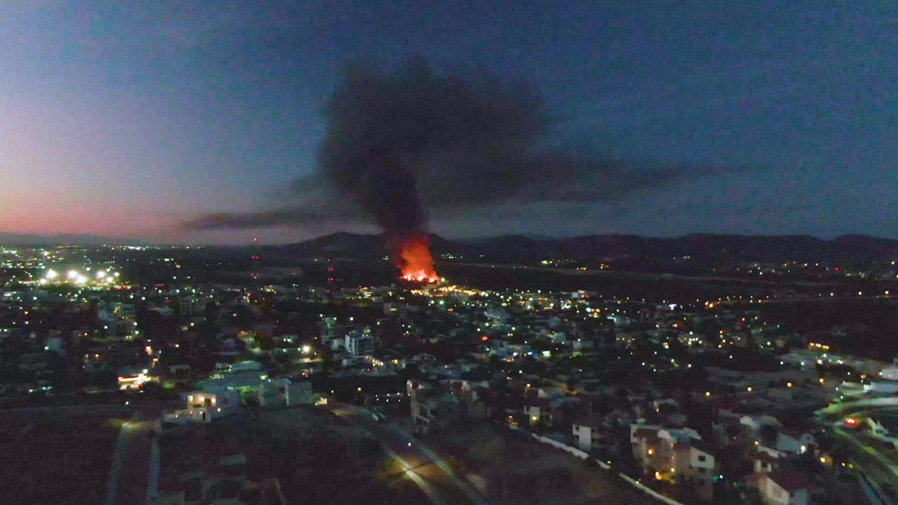 Aerial view of San Jose del Cabo at dusk with dense city lights below, a bright fire in the distance, black smoke drifting across the sky, and no visible people.
