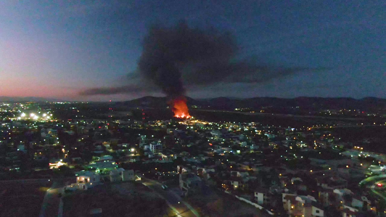 Wide aerial frame over San Jose del Cabo showing lit streets and buildings in the foreground, orange flames near the horizon, a broad black smoke cloud, and no visible people.