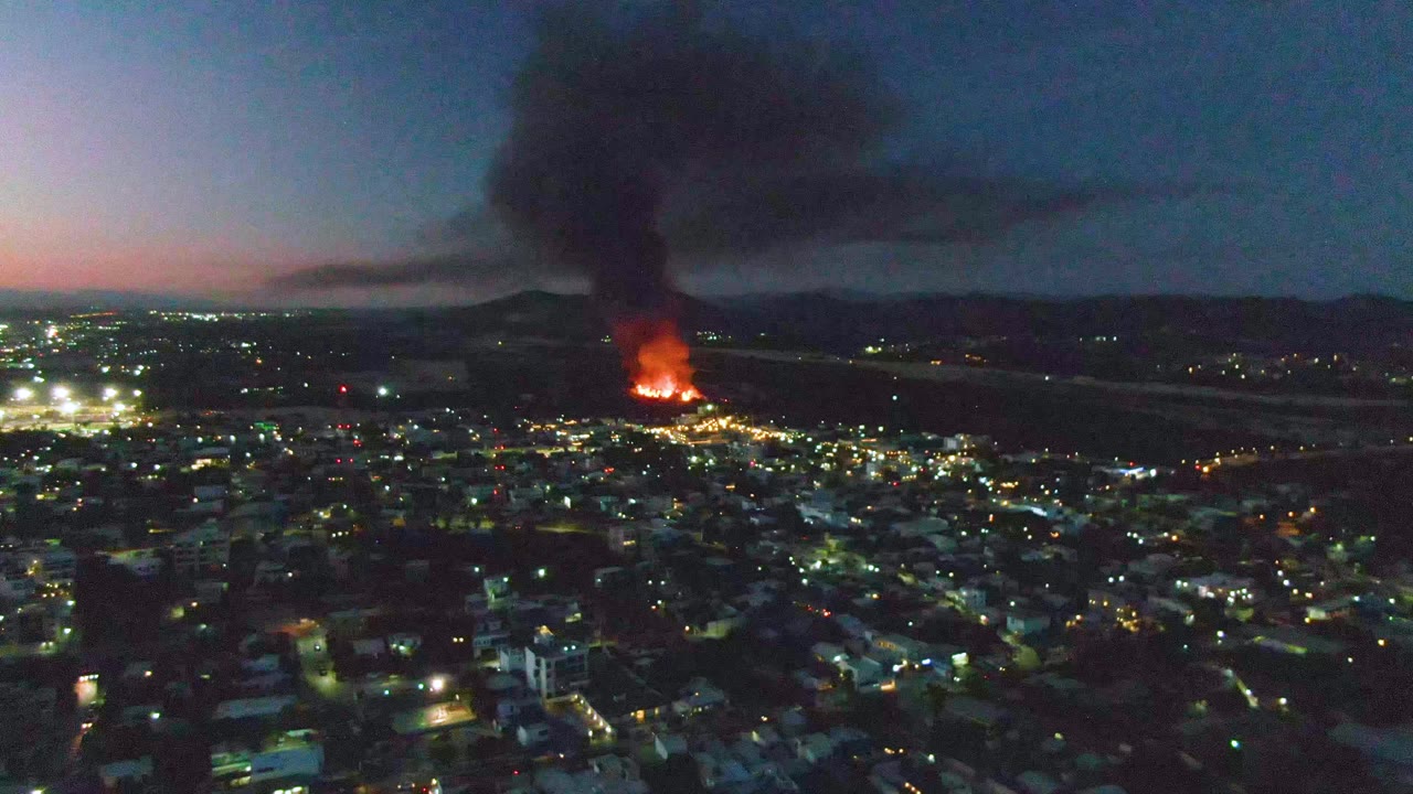 A wide aerial view over San Jose del Cabo at dusk shows city lights below, orange flames in the distance, a black smoke column above, and no visible people.