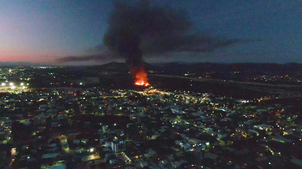 San Jose del Cabo appears from a high aerial angle with a bright fire near the horizon, scattered street lights below, black smoke spreading overhead, and no visible people.