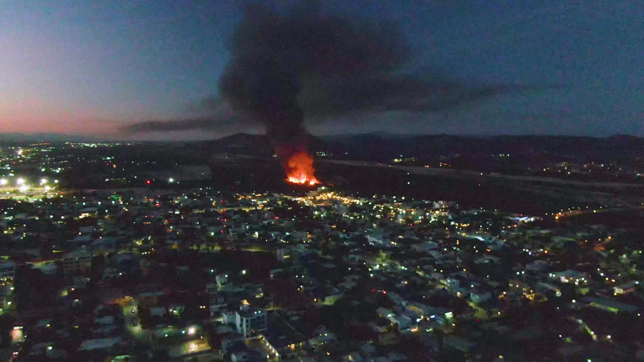 A broad San Jose del Cabo cityscape shows clustered lights in the foreground, a concentrated orange blaze in the distance, dense black smoke, and no visible people.